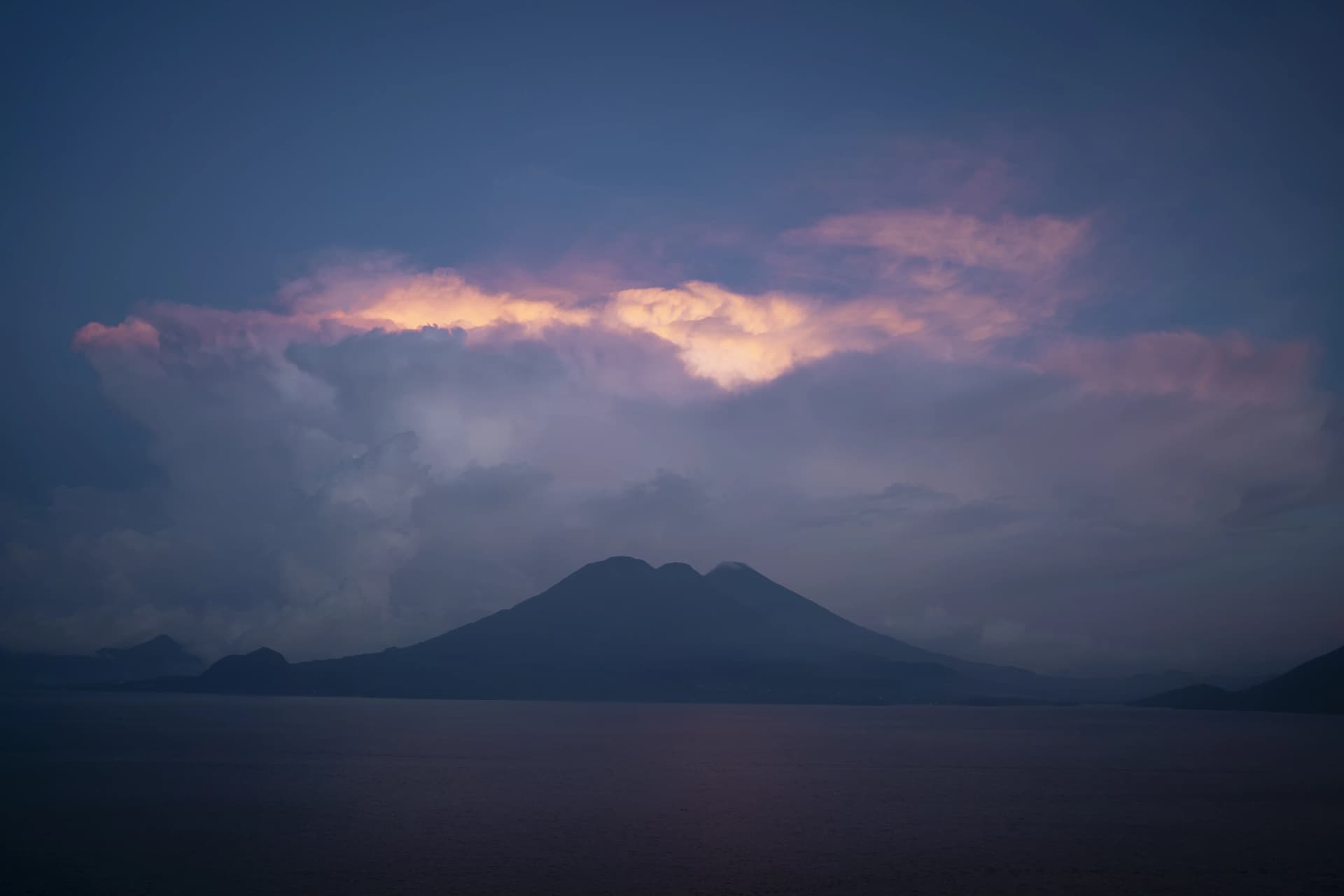 Twin-peaked volcano silhouette across Lake Atitlán at dusk, storm clouds catching the last pink light